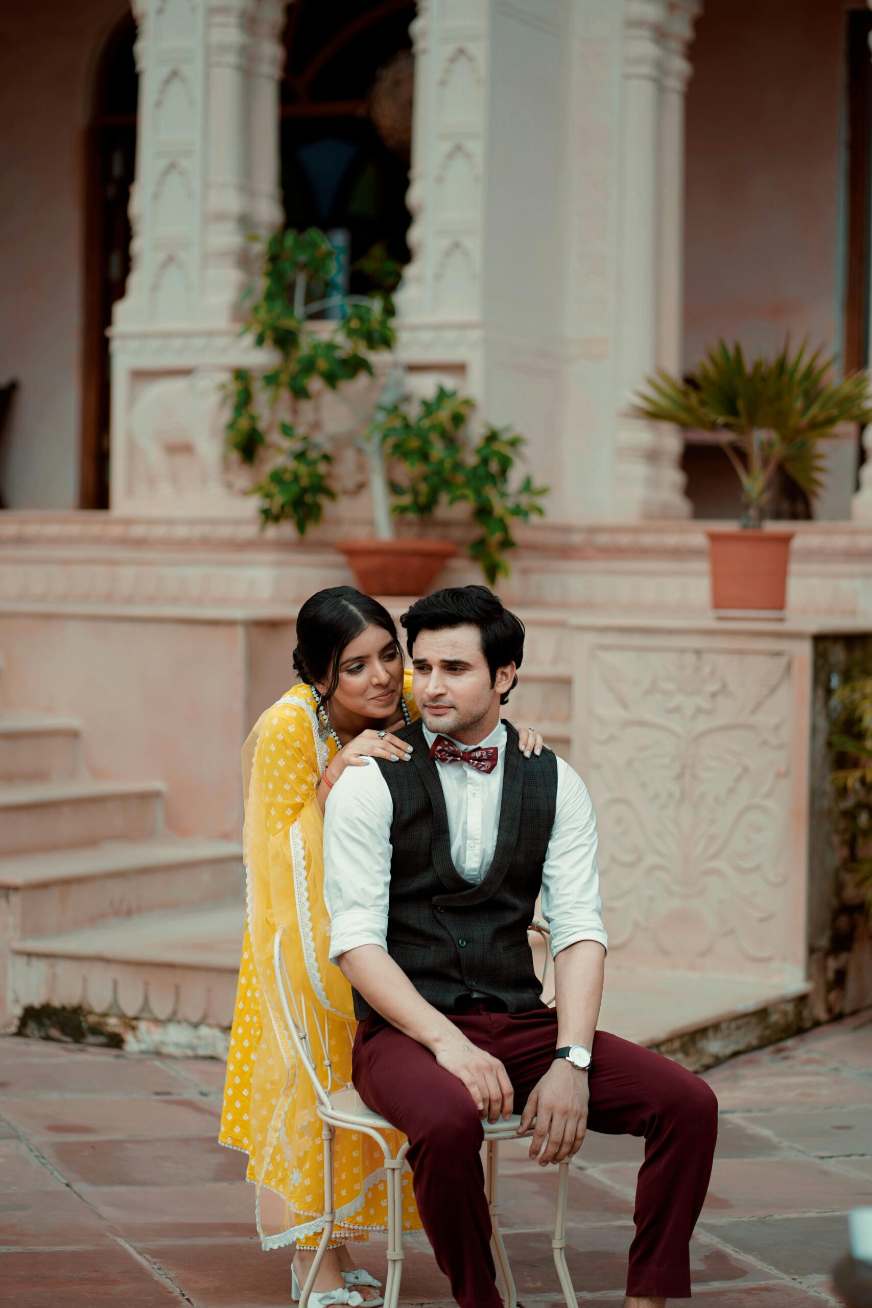 A vibrant South Asian couple in elegant attire posing outdoors with artistic architecture.